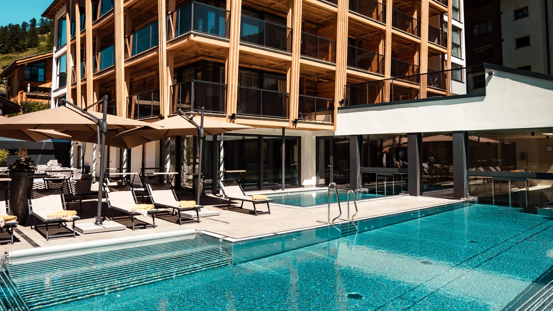 Resort La Ginabelle Zermatt: Ihr 4*S Wellnessresort in Zermatt Gebäude im Sommer mit grossem Aussenpool und Ruheliegen.